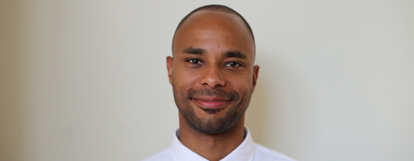 Image contains a photo of a man smiling at the camera. The man is wearing a white dress shirt and standing in front of a white background.